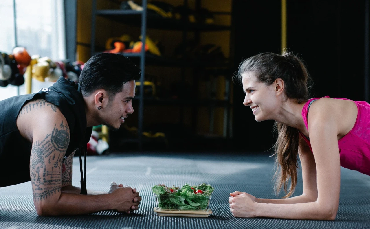 Dos personas hacen planchas frente a frente en el gimnasio con una ensalada, representando la relación entre alimentación equilibrada y ejercicio físico